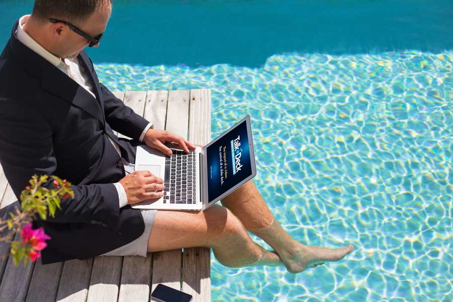 Man in suit jacket with bare legs working on a laptop by the pool; screen shows Talk-Deck tagline about presentation engagement.
